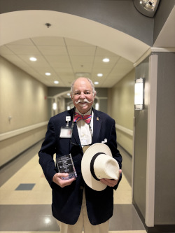 Man stands in hallway holding plaque, smiling